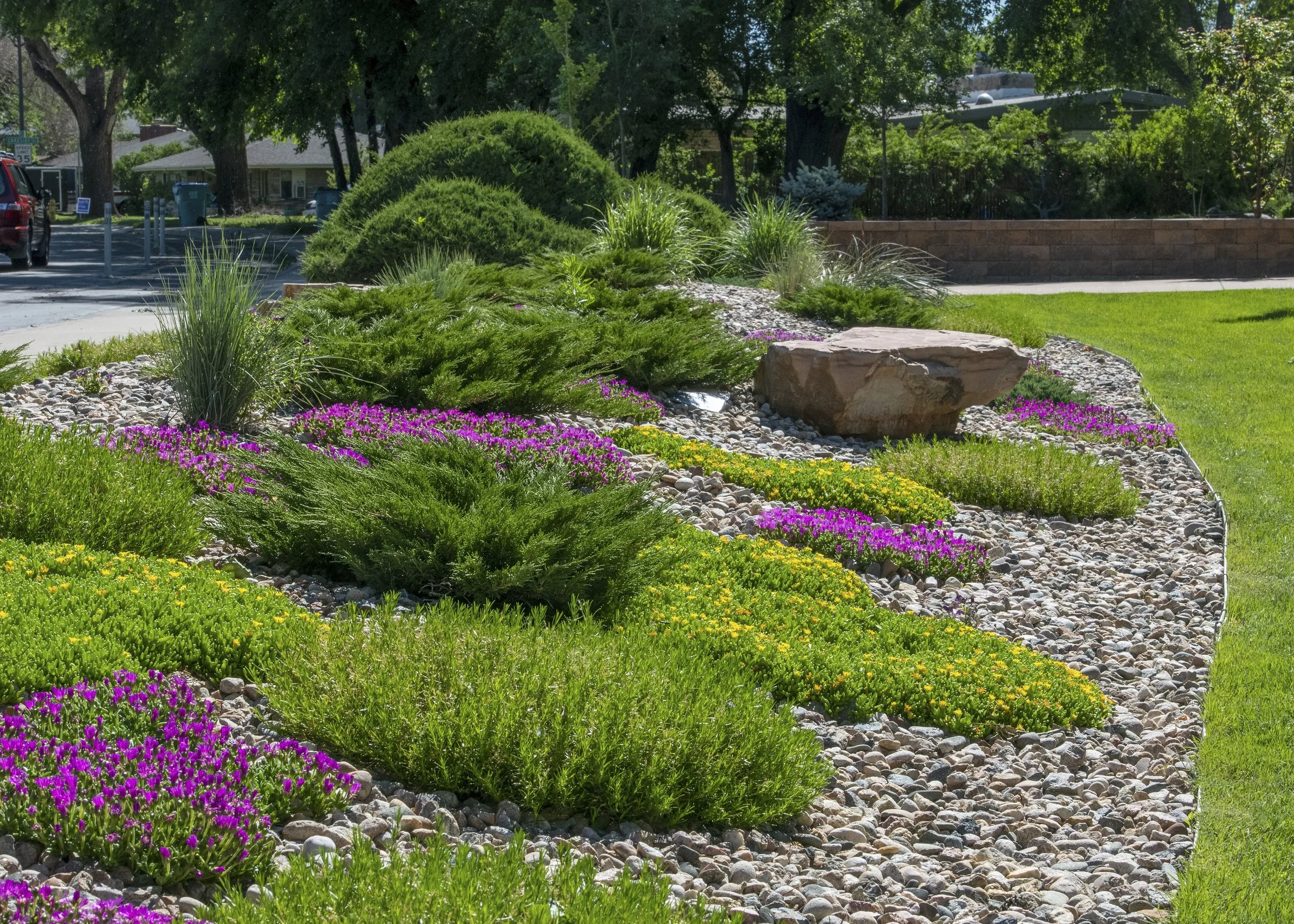 A landscaped garden with colorful flowers, green bushes, and rocks, next to a grassy area with trees and a sidewalk, in a sunny outdoor setting.