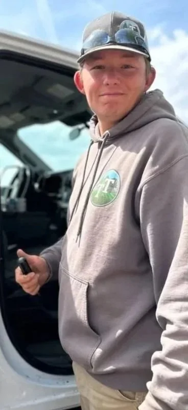 Young man standing outdoors in front of a vehicle, wearing a gray hoodie with a logo, a cap, and sunglasses on top of the cap, holding a remote or a small device in his right hand.
