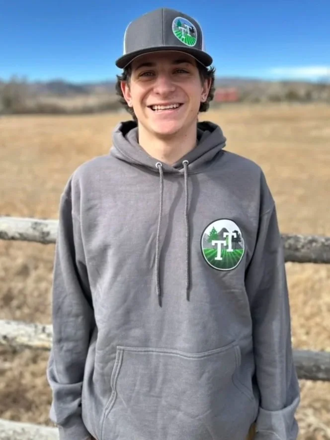 A young man standing outdoors in a rural area, smiling, wearing a gray hoodie and a gray baseball cap with a green and white logo. The hoodie and cap both have a matching logo featuring trees, a mill, and the letter T.