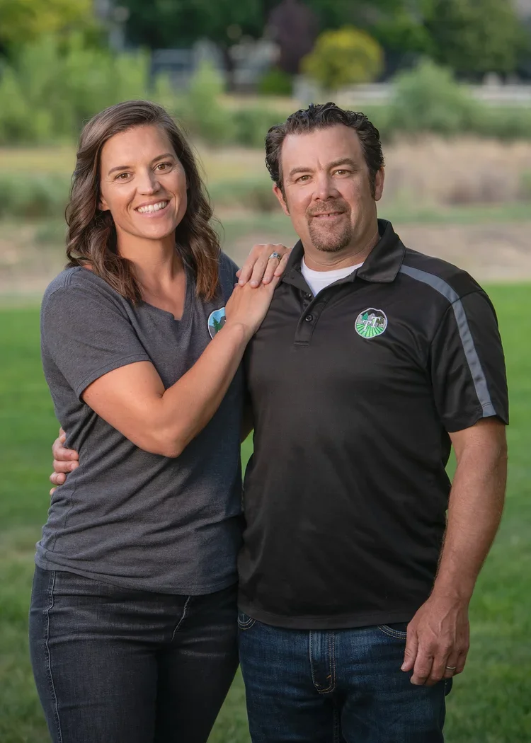 A smiling woman and man stand close together outdoors, with the woman resting her hand on the man's shoulder in a park with green grass and trees in the background.
