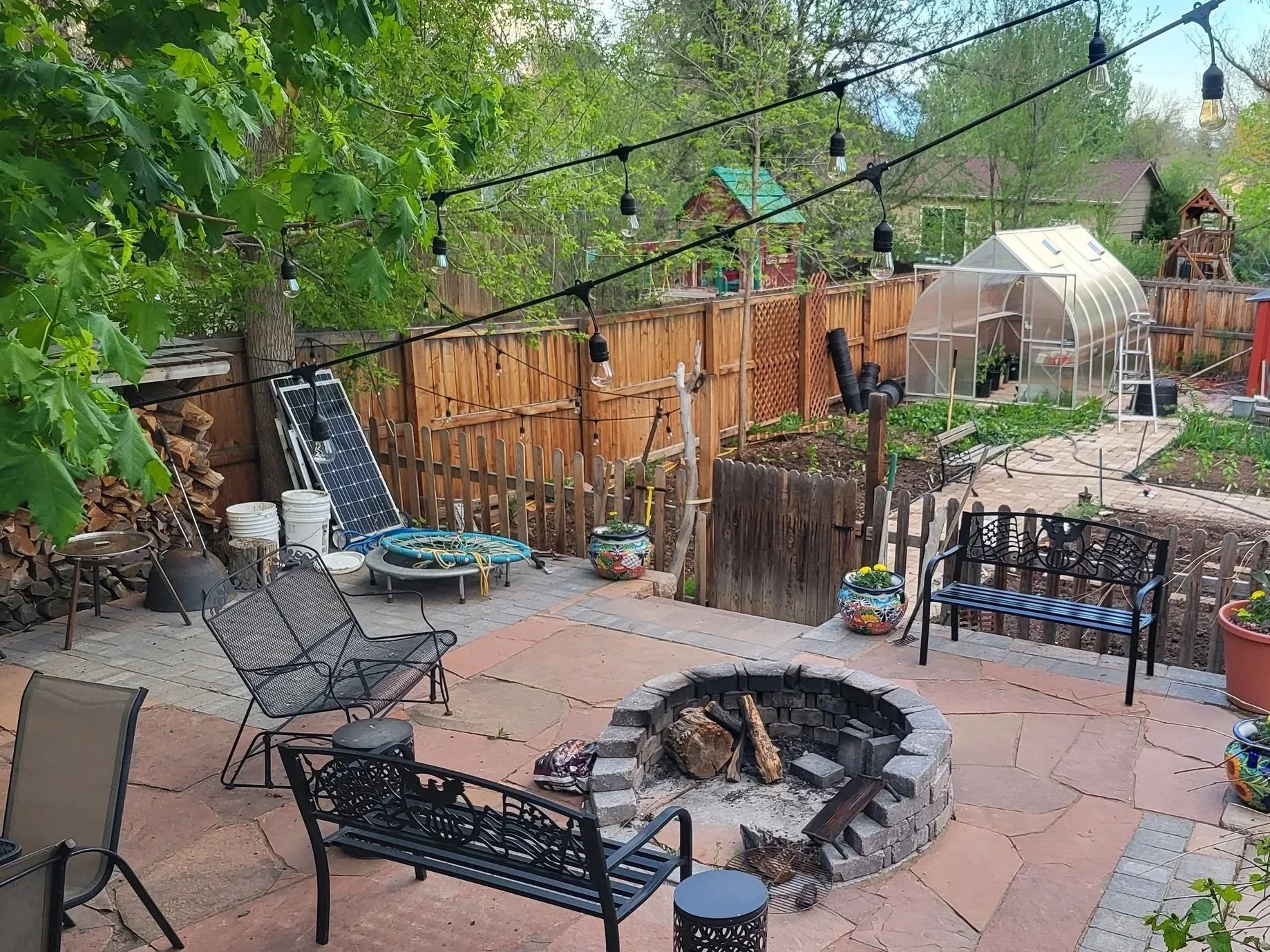 Backyard patio with a fire pit, metal and wood chairs, potted plants, string lights, a solar panel, a greenhouse, and a fenced garden area surrounded by trees and a wooden fence.