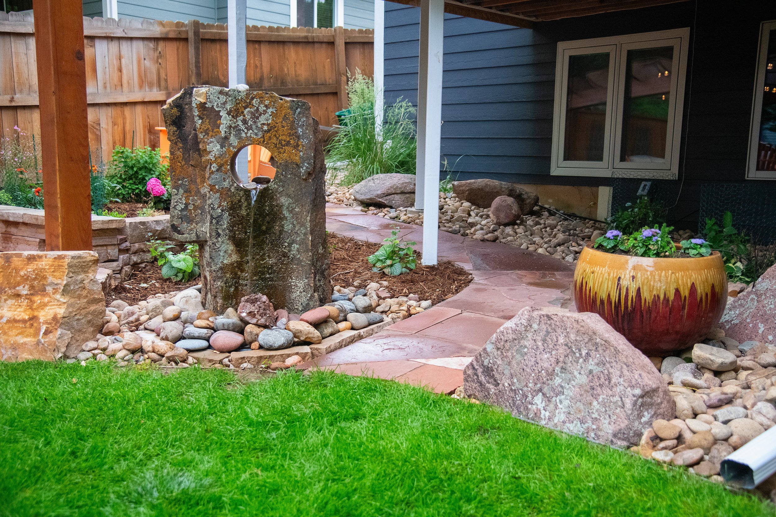 A backyard garden with a stone fountain that has a circular hole at the top, surrounded by decorative rocks and small plants. There is a curved red stone pathway, a colorful ceramic planter with purple flowers, and a dark blue house with windows in t