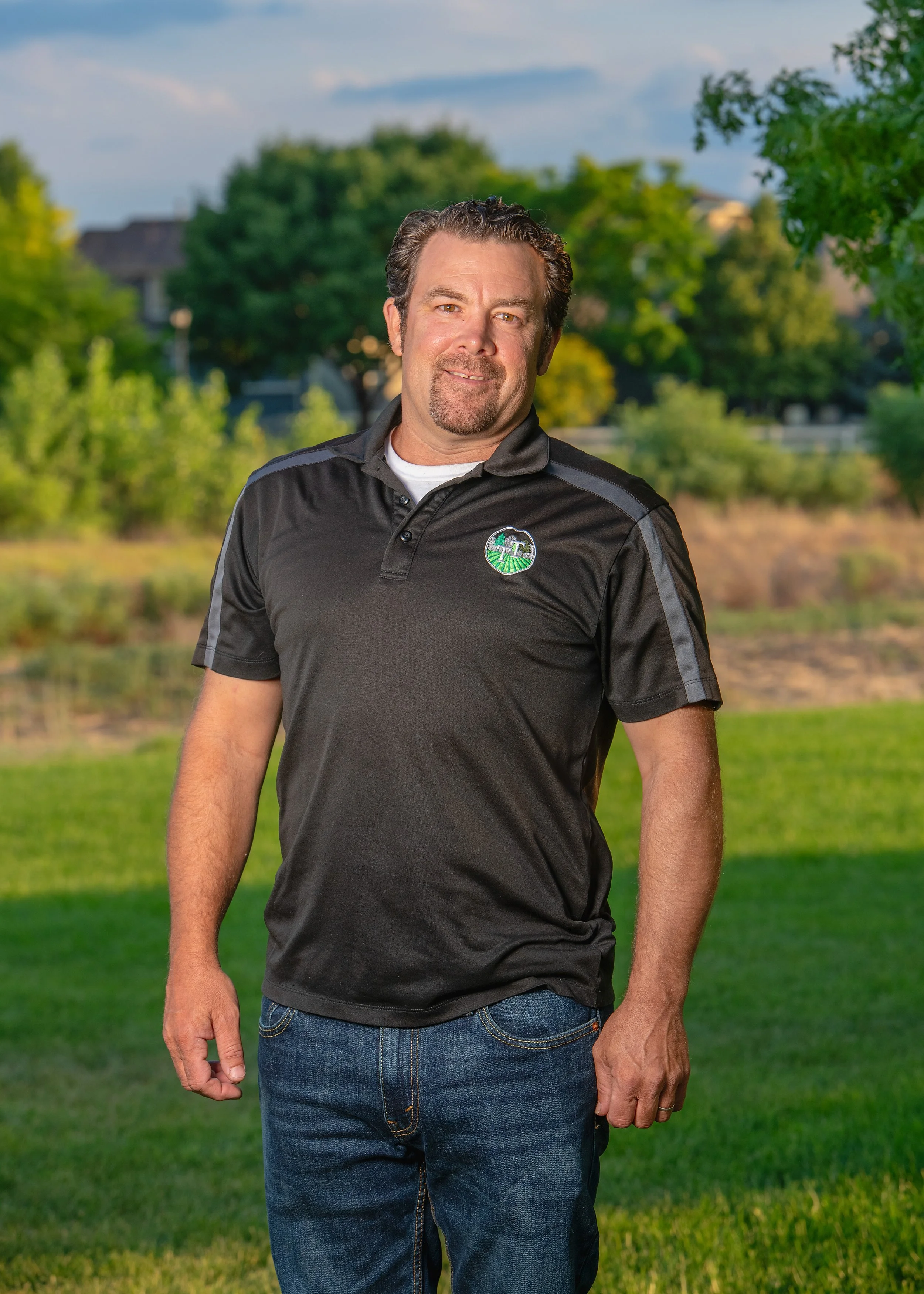 A man with dark wavy hair and a goatee, wearing a black collared shirt with a green and white logo, stands outdoors on a grassy area with trees and houses in the background. He is smiling and looking at the camera.