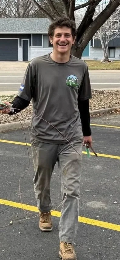 Smiling man walking in parking lot holding a wire and a pair of pliers, dressed in gray outdoor work clothes, with leafless trees and residential houses in the background.