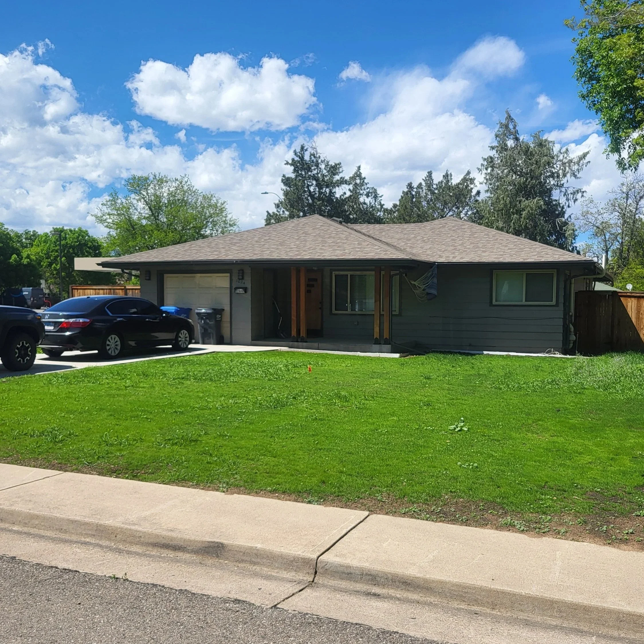 Single-story house with a gray exterior, brown roof, and a three-car garage. Two black cars parked in the driveway. Green lawn in front, trees and blue sky with clouds in the background.