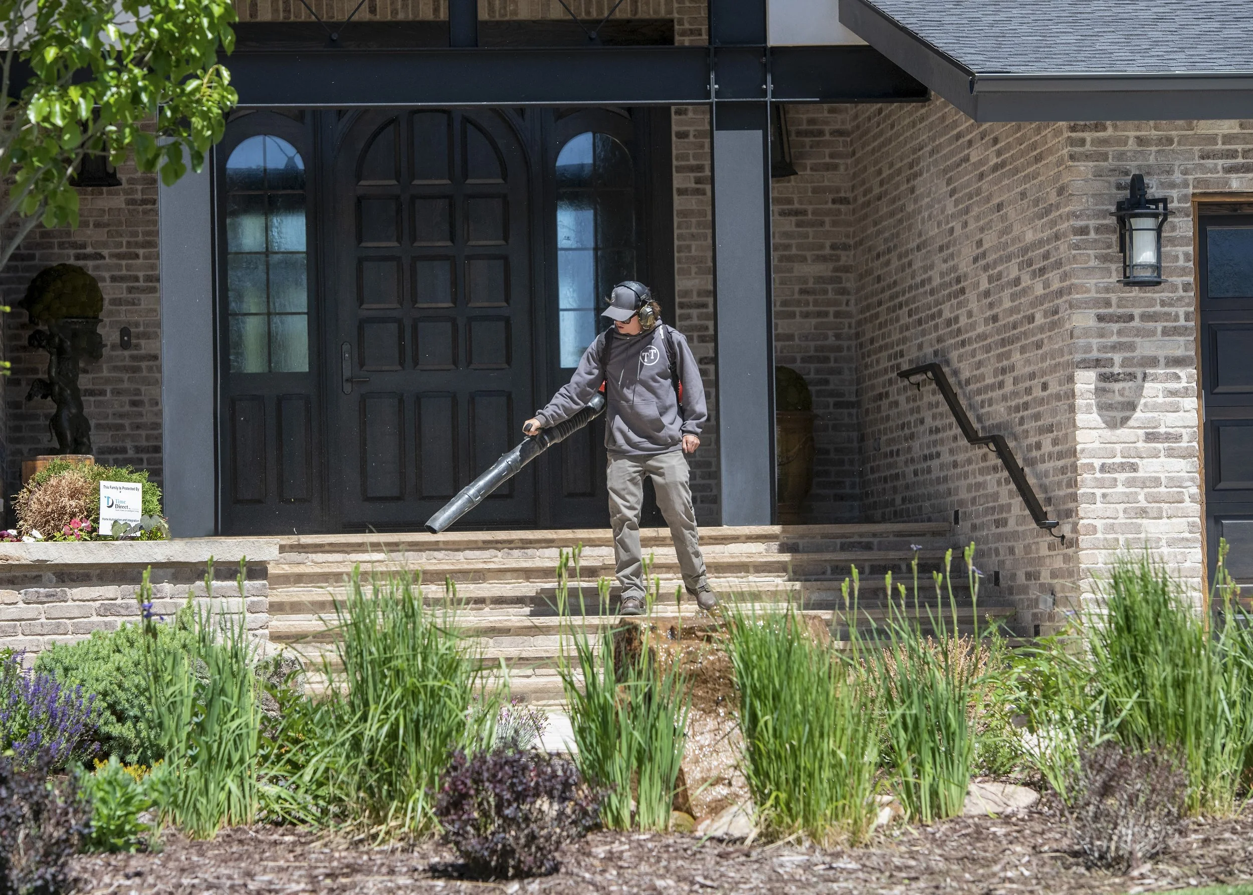 A person wearing a hoodie, cap, and headphones is using a leaf blower to clean the front steps of a brick house with a black door and exterior light fixture.