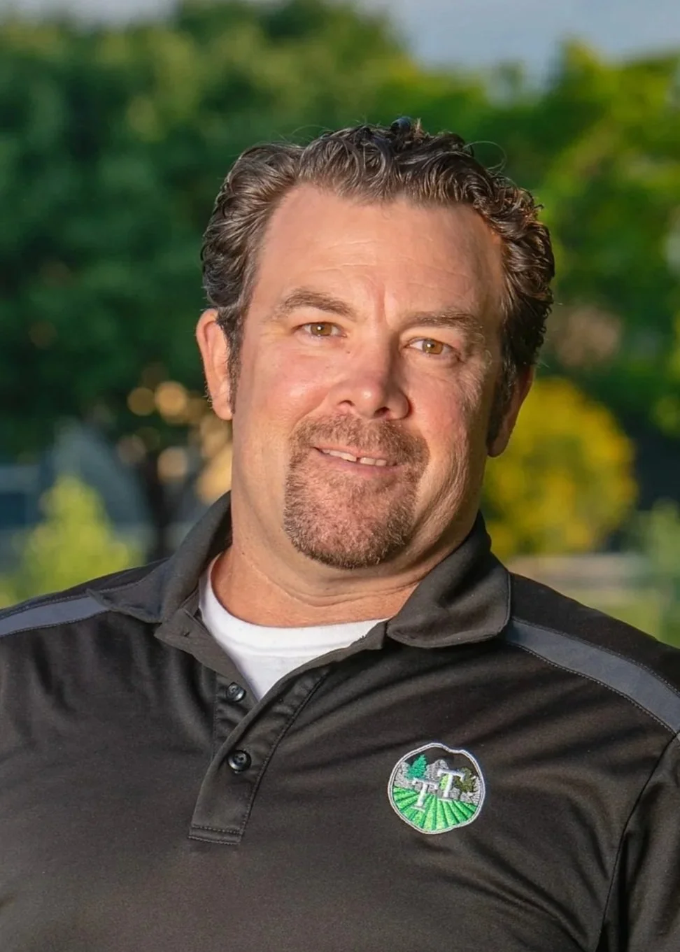 A man with brown hair and a beard smiling outdoors in front of green trees, wearing a black collared shirt with a logo of a farm scene.