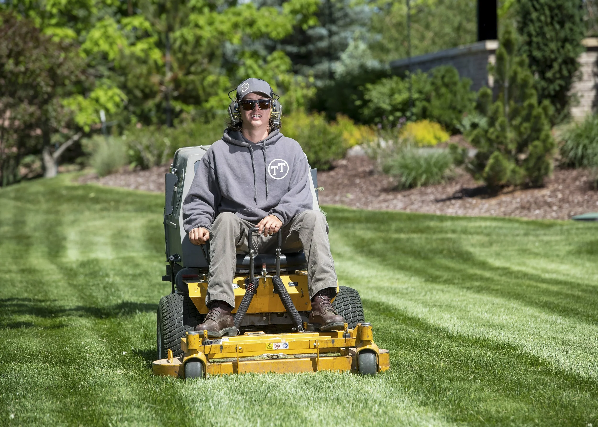 Person mowing a lawn with a riding lawn mower in a garden with green bushes and trees.