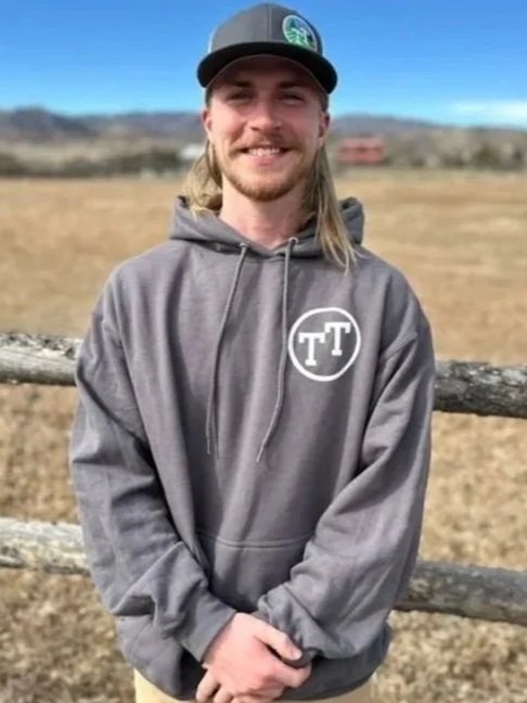 Young man with long blonde hair, gray hoodie with white logo, and gray baseball cap, standing outdoors near a wooden fence with open field and blue sky in the background.