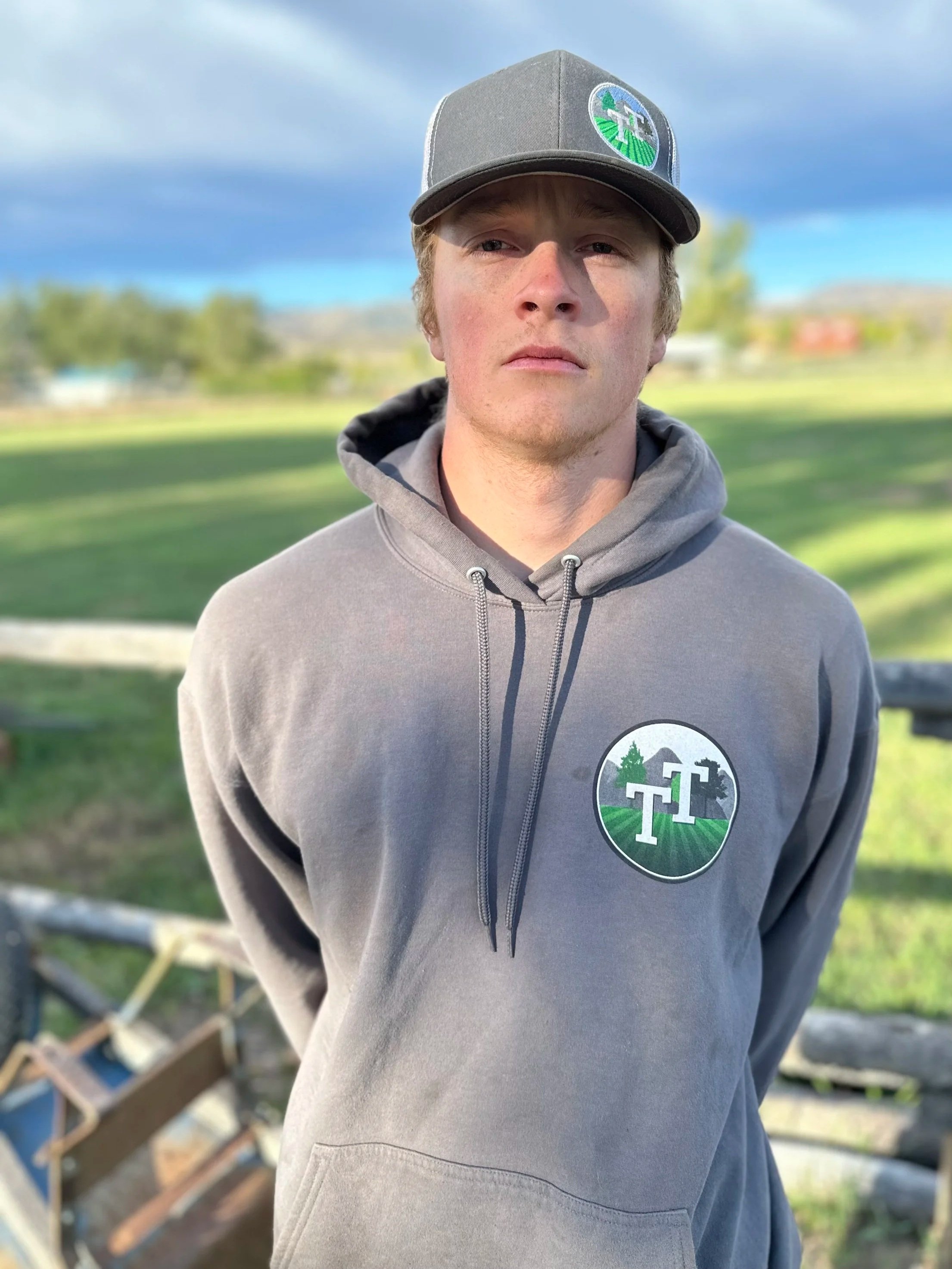 Young man wearing a gray hoodie and matching cap with a logo, standing outdoors in a park with a blurred background of trees, grass, and a wooden fence.