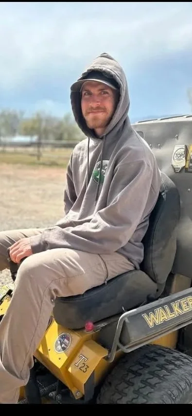 A man wearing a gray hoodie sitting on a yellow golf cart outdoors with a cloudy sky in the background.