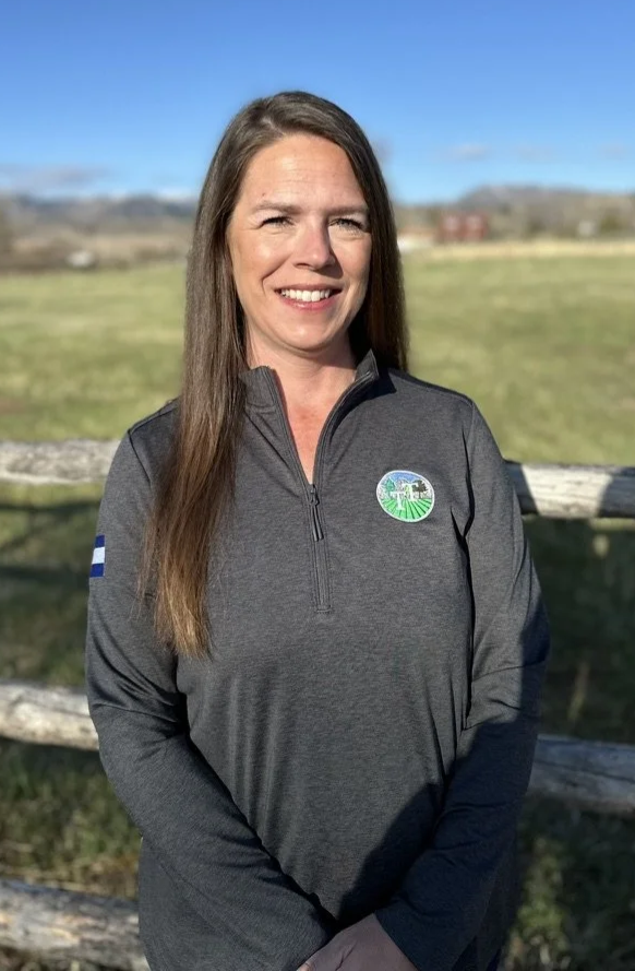 A woman standing outdoors in front of a wooden fence with open fields and hills in the background. She is smiling, has long brown hair, and is wearing a gray quarter-zip jacket with a logo on the chest and an American flag patch on the sleeve.