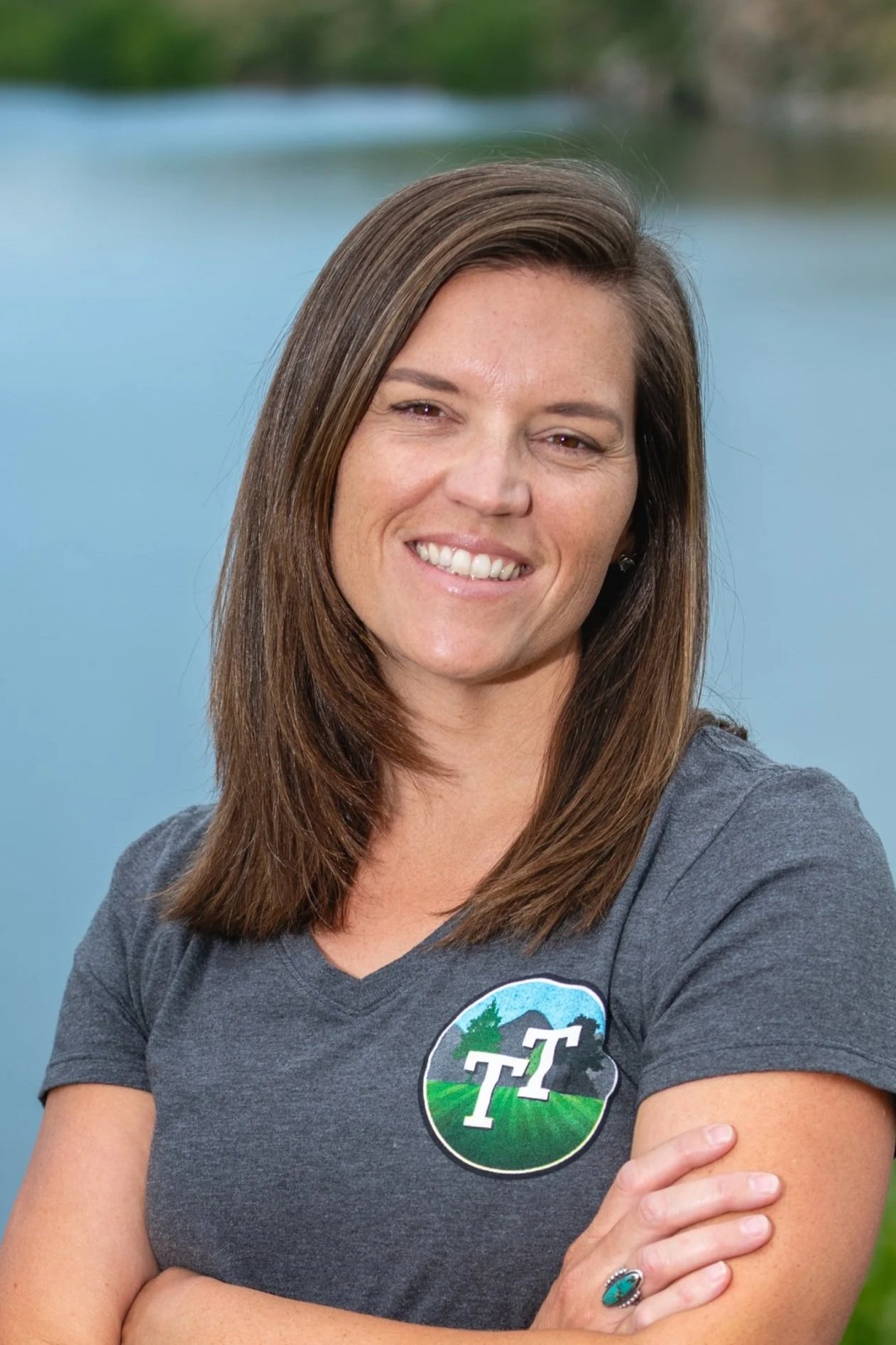A woman with shoulder-length brown hair smiling with arms crossed, wearing a gray T-shirt with a logo, standing outdoors with a blurred blue sky and trees in the background.