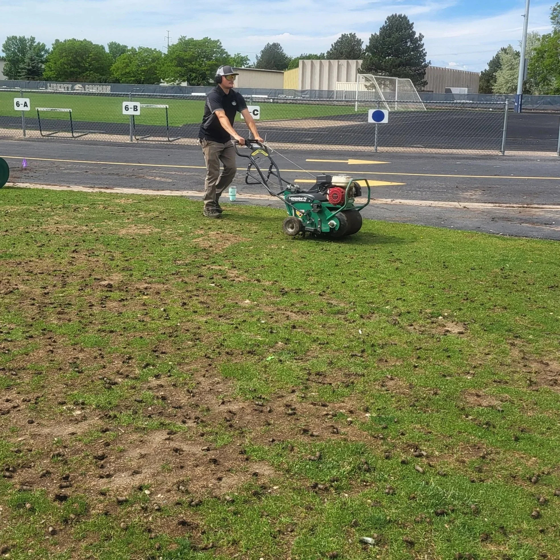 A man is using a power roller to aerate or fertilize a grassy field near a track and football field at a school or sports complex.