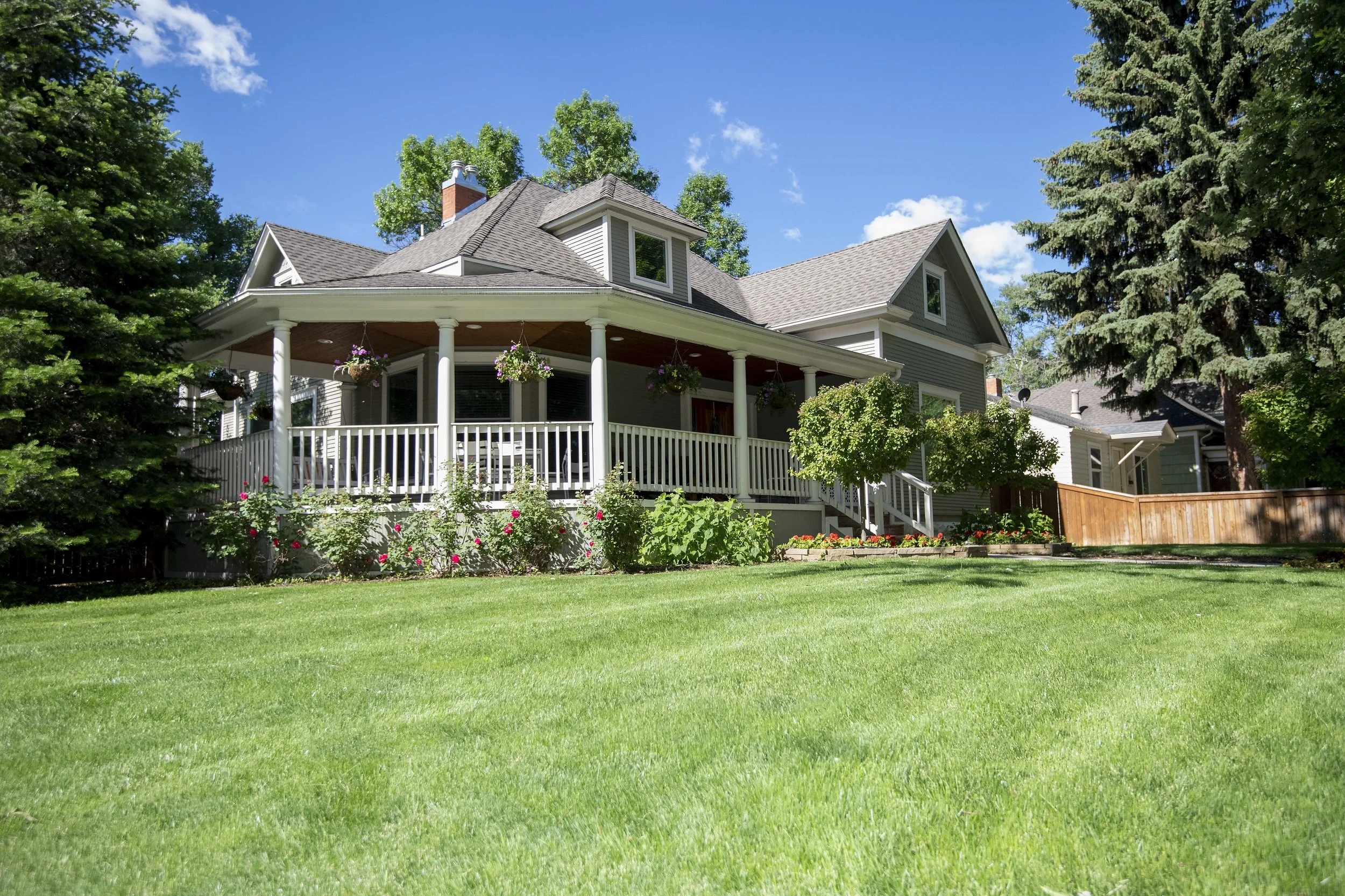 A large house with a wrap-around porch, multiple gabled roofs, and a well-maintained lawn with bushes and trees in a suburban neighborhood under a clear blue sky.