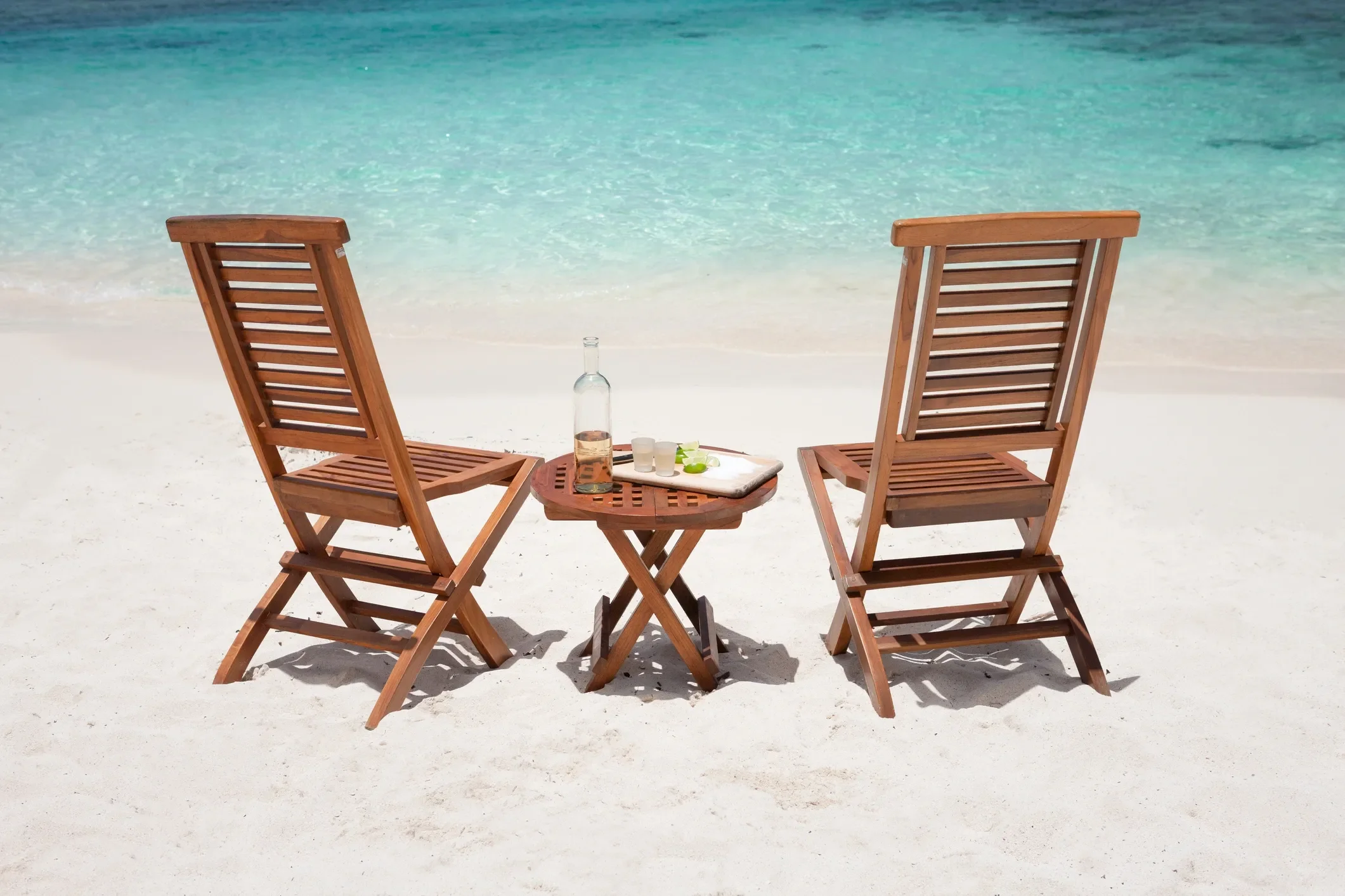 Two wooden lounge chairs on a sandy beach with a small wooden table between them, holding a bottle, glasses, and lime slices, with clear turquoise ocean water in the background.