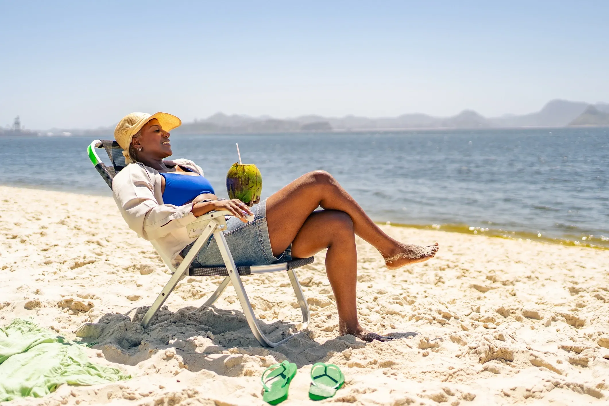 A woman relaxing on a beach chair, wearing a yellow hat, blue swimsuit, and denim shorts. She is smiling, holding a coconut drink with a straw, and has her legs crossed. There are green flip-flops on the sand nearby, with the ocean and mountains in the background.