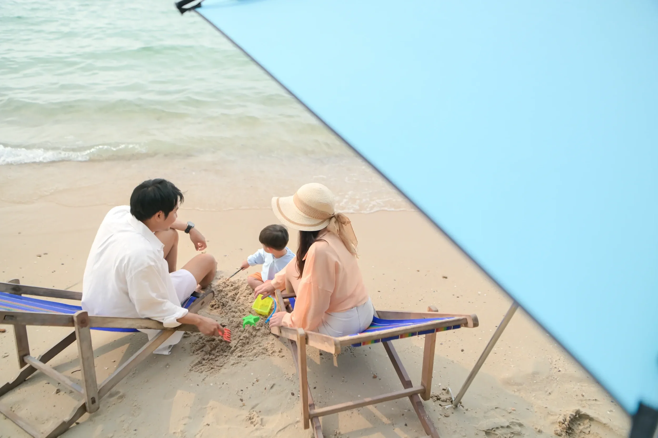 A family building a sandcastle on the beach under a blue umbrella, with the ocean in the background.