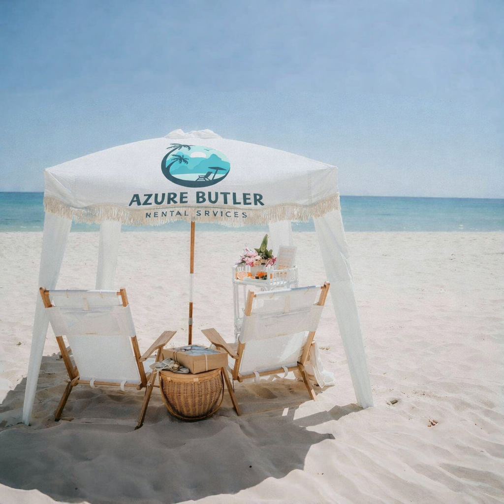 A beach cabana with two lounge chairs, a small table with flowers, under a white canopy that reads 'Azure Butler Rental Services'. The sand is white, and the ocean is blue with a clear sky.