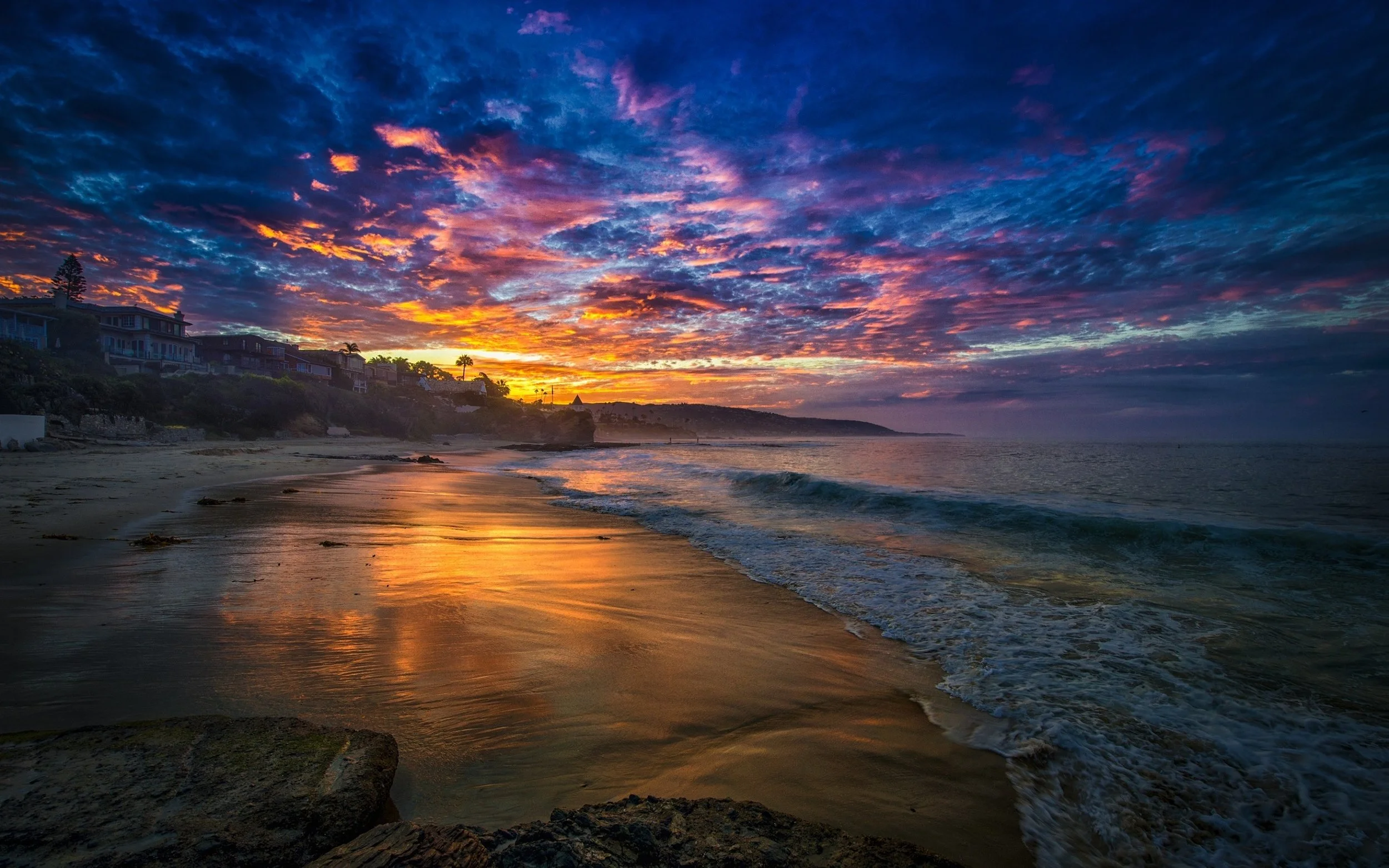Colorful sunset over a beach with houses on a hillside, reflecting on wet sand and gentle waves.