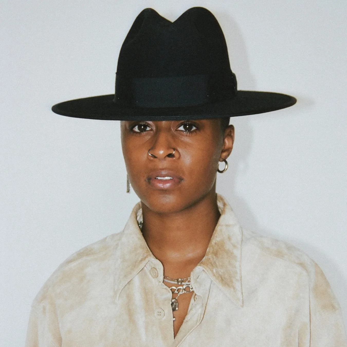A portrait of a woman wearing a large black hat, beige shirt, and layered silver necklaces, with a neutral background.