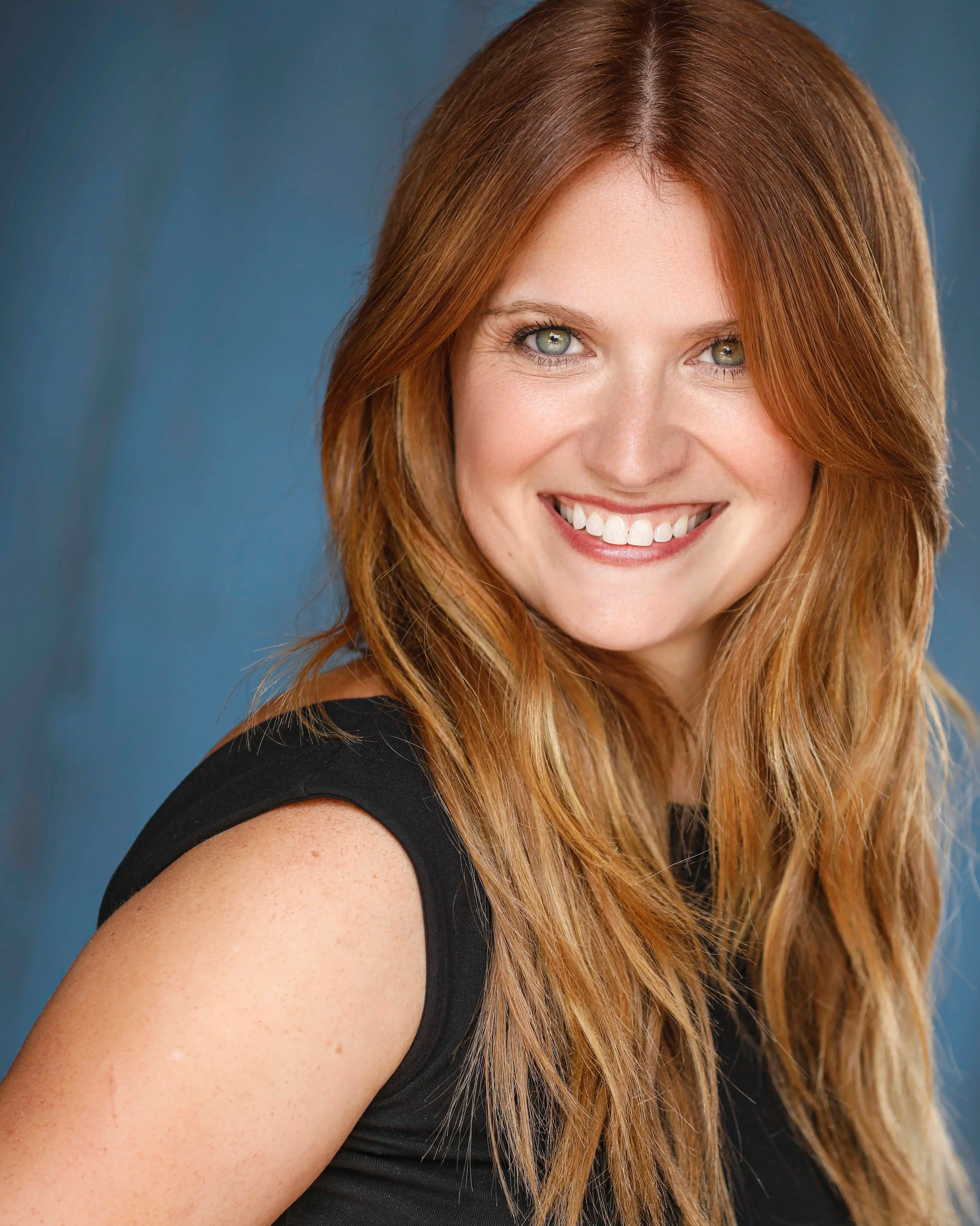 Close-up of a smiling woman with long red hair, wearing a black sleeveless top, against a blue background.