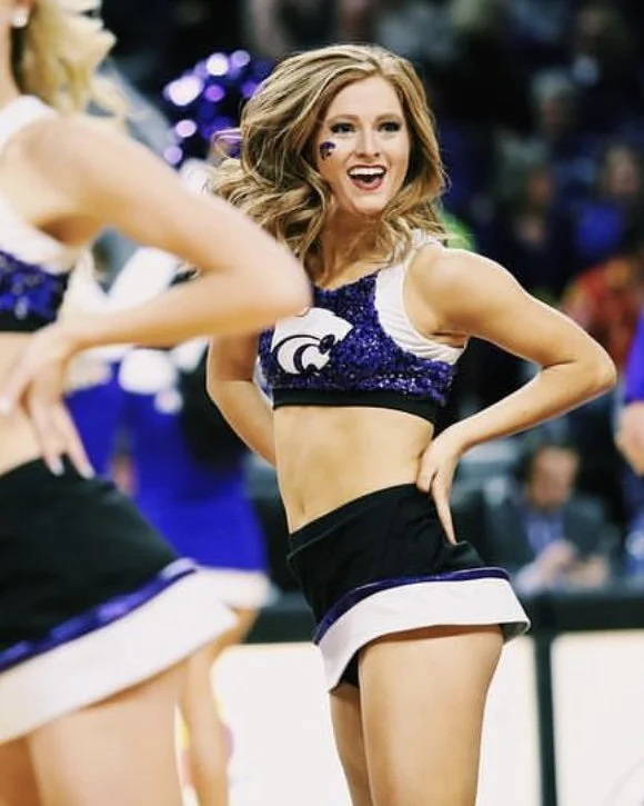 Cheerleader with long, wavy blonde hair, smiling, wearing a purple and white crop top and black cheerleading skirt, standing with one hand on her hip during a cheerleading performance.
