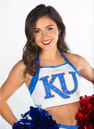 Young woman dressed as a cheerleader in blue and white with 'KU' on her top, holding pom-poms, smiling.