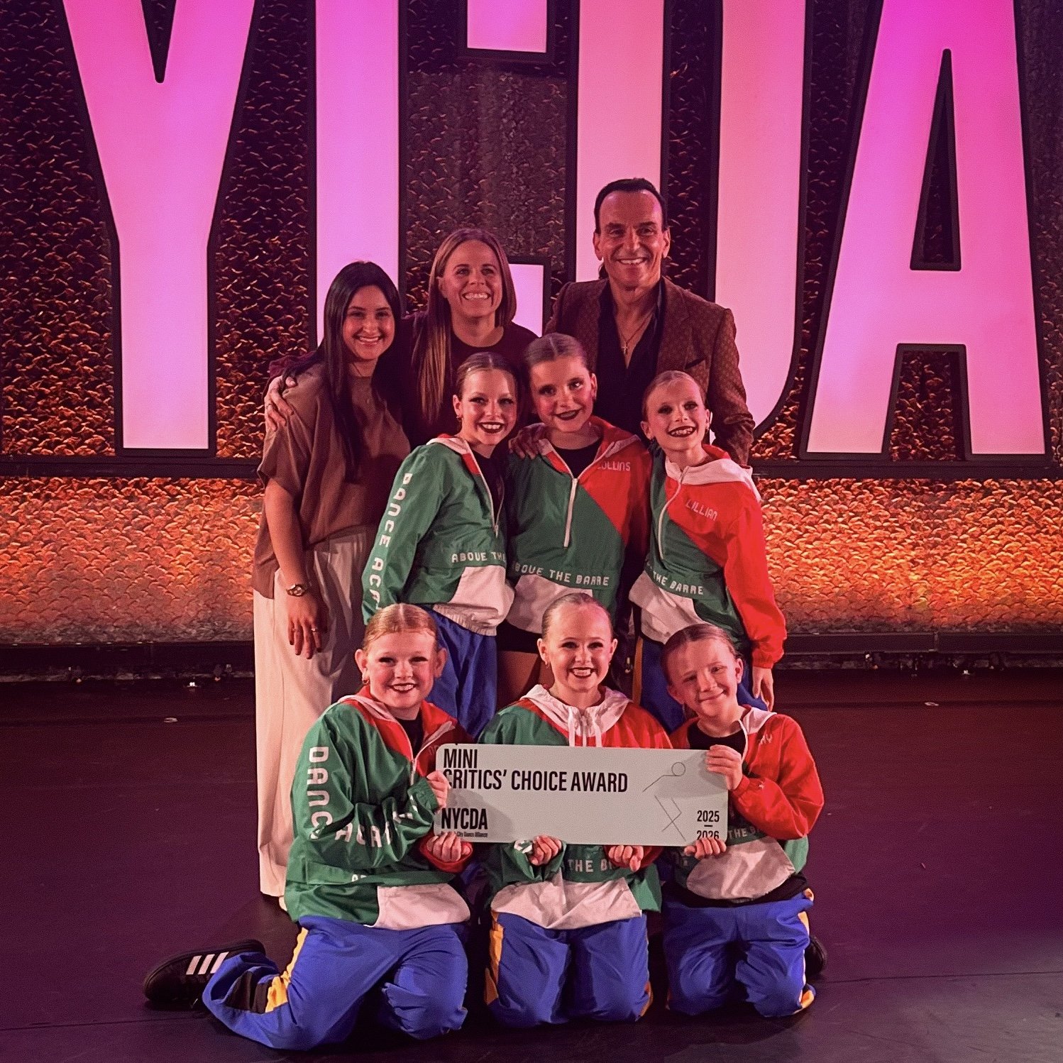 A group of young girls in colorful jackets holding an award plaque with the text 'Mini Critics' Choice Award', posing on a stage with a large illuminated sign in the background.