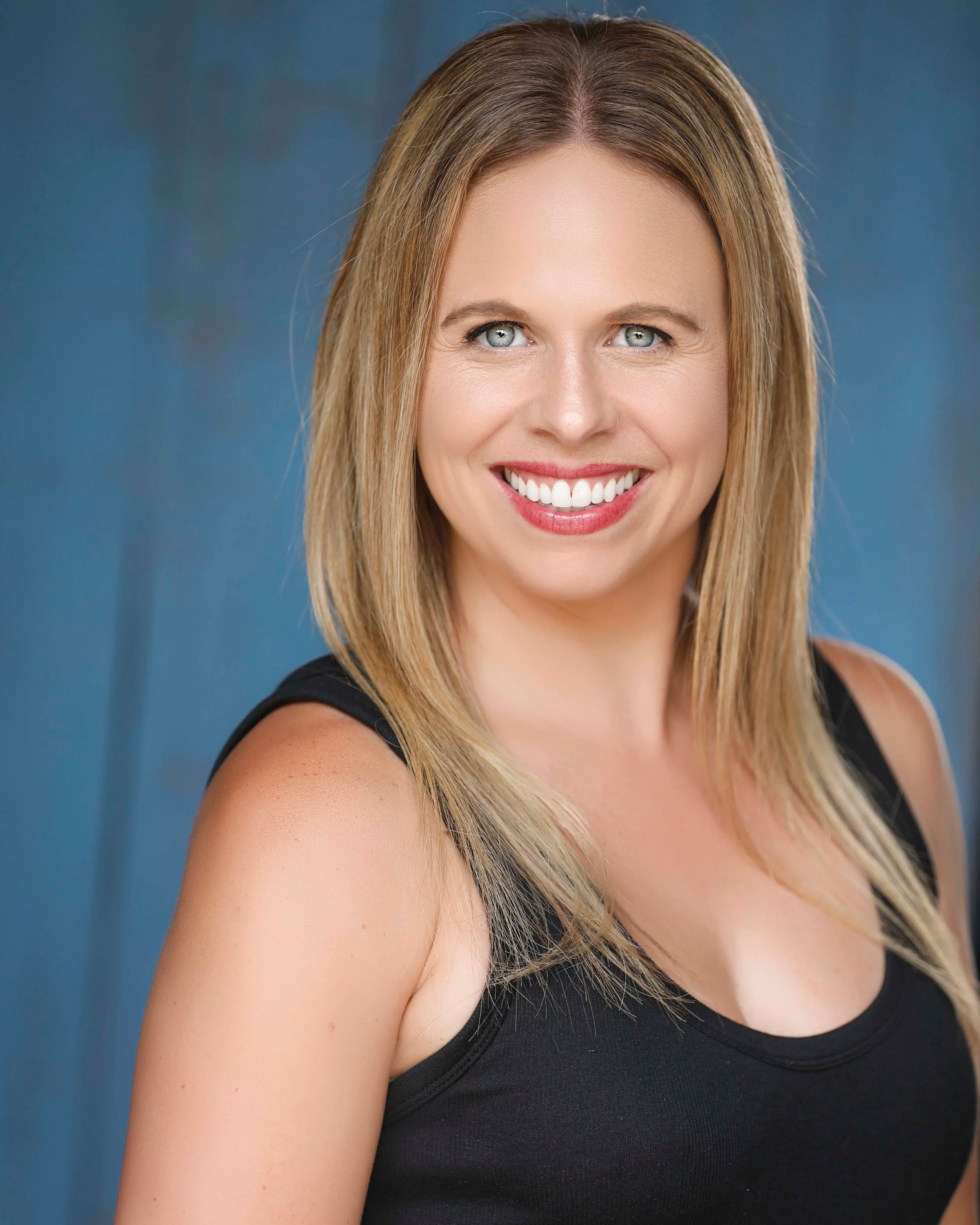 A woman with blonde hair, blue eyes, and fair skin, smiling, wearing a black sleeveless top, with a blurred blue background.