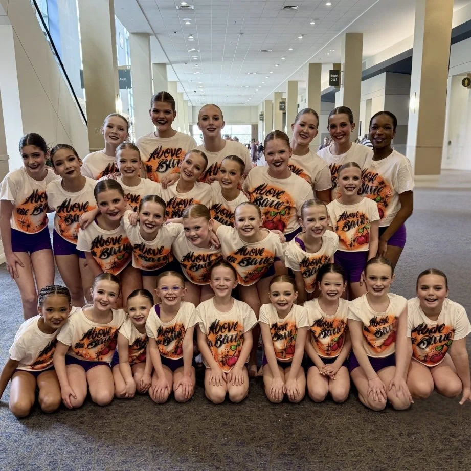 Group of young girls in matching dance team outfits posing together indoors at a convention or competition.