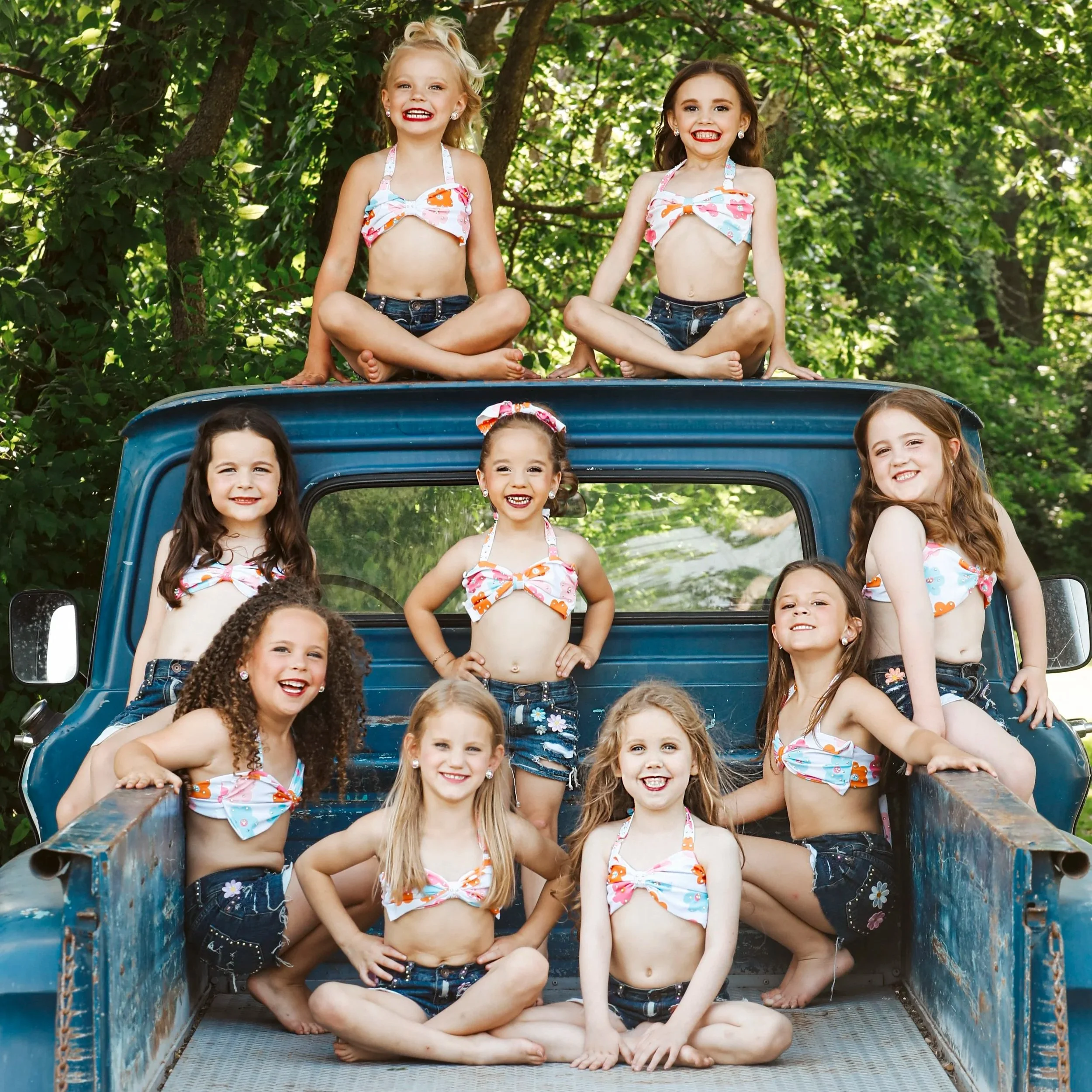 Group of ten young girls dressed in matching outfits with floral tops, denim shorts, and headbands standing in front of a curtain.