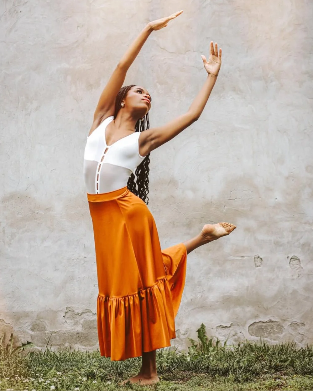 Woman in an orange skirt and white top performing a dance pose against a plain wall.