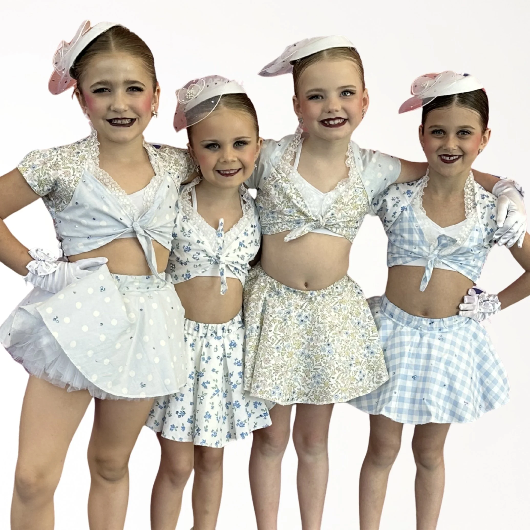 Four young girls dressed in vintage-style outfits with skirts and blouses, wearing matching hats, posing with their arms around each other.