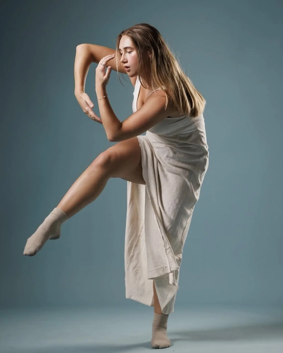 A young woman with long hair is posing against a plain background. She is wearing a beige dress and socks, and she is striking a dance pose with one leg raised and her eyes closed.