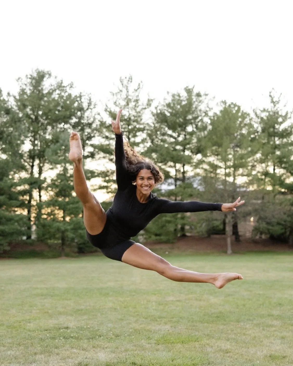 A woman in black athletic wear performing a leap outdoors on a grassy field with trees in the background.