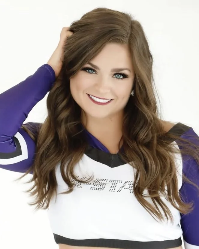 A young woman with long brown hair, wearing a purple and black sports jersey, smiling and touching her hair.