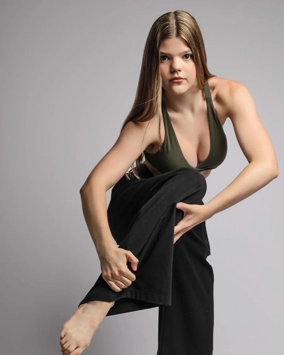 A young woman with long light brown hair, wearing a green tank top and black pants, is in a pose where she is holding her left foot with her left hand and looking directly at the camera against a plain gray background.