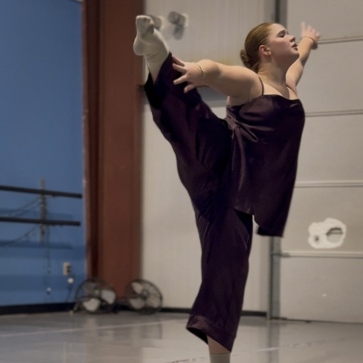 Young woman practicing ballet in a garage, wearing a dark dress and white ballet shoes, holding her leg in a high arabesque pose.