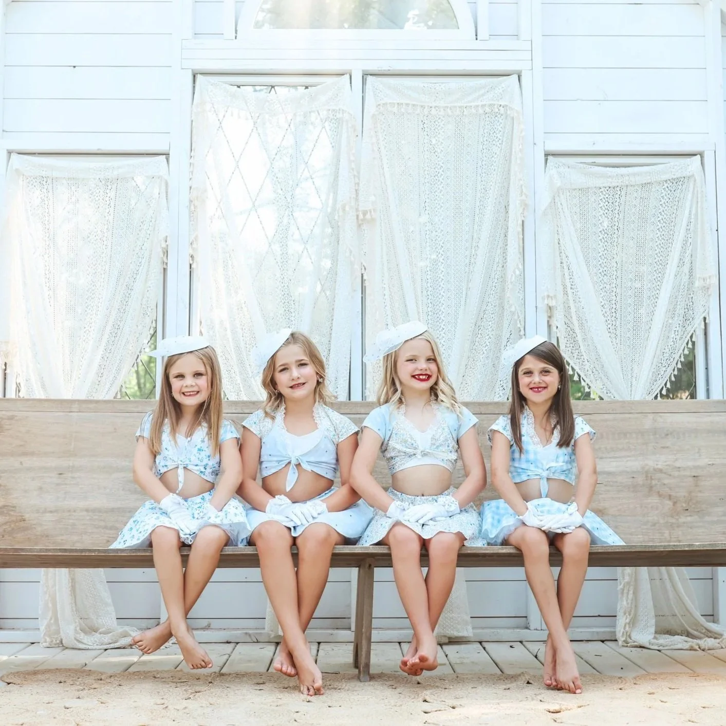 Four young girls dressed in vintage-style outfits with skirts and blouses, wearing matching hats, posing with their arms around each other.