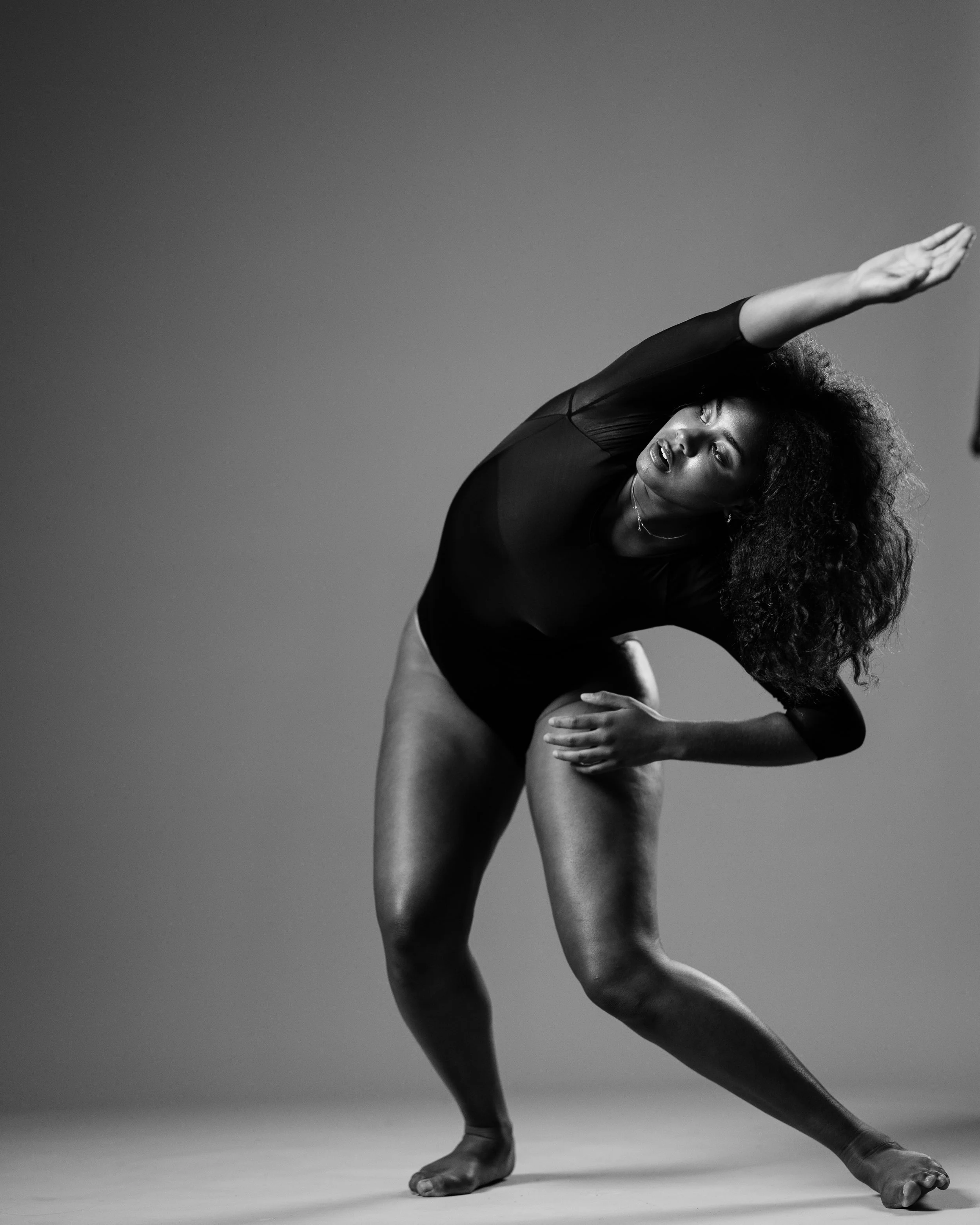 Black and white photo of a woman with curly hair in a dance pose, with one arm extended overhead and her body slightly bent sideways.
