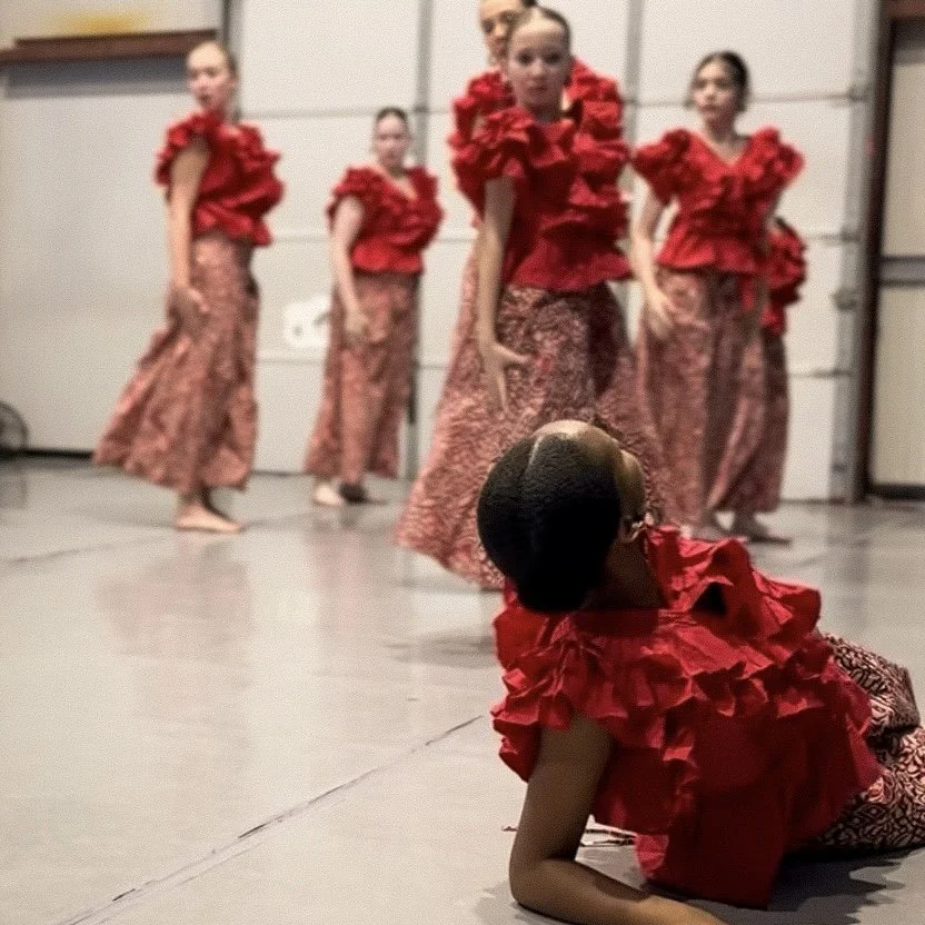 A group of five women in matching red ruffled tops and patterned skirts standing in a room, with one woman in the foreground lying on the floor, also wearing a matching red ruffled top.