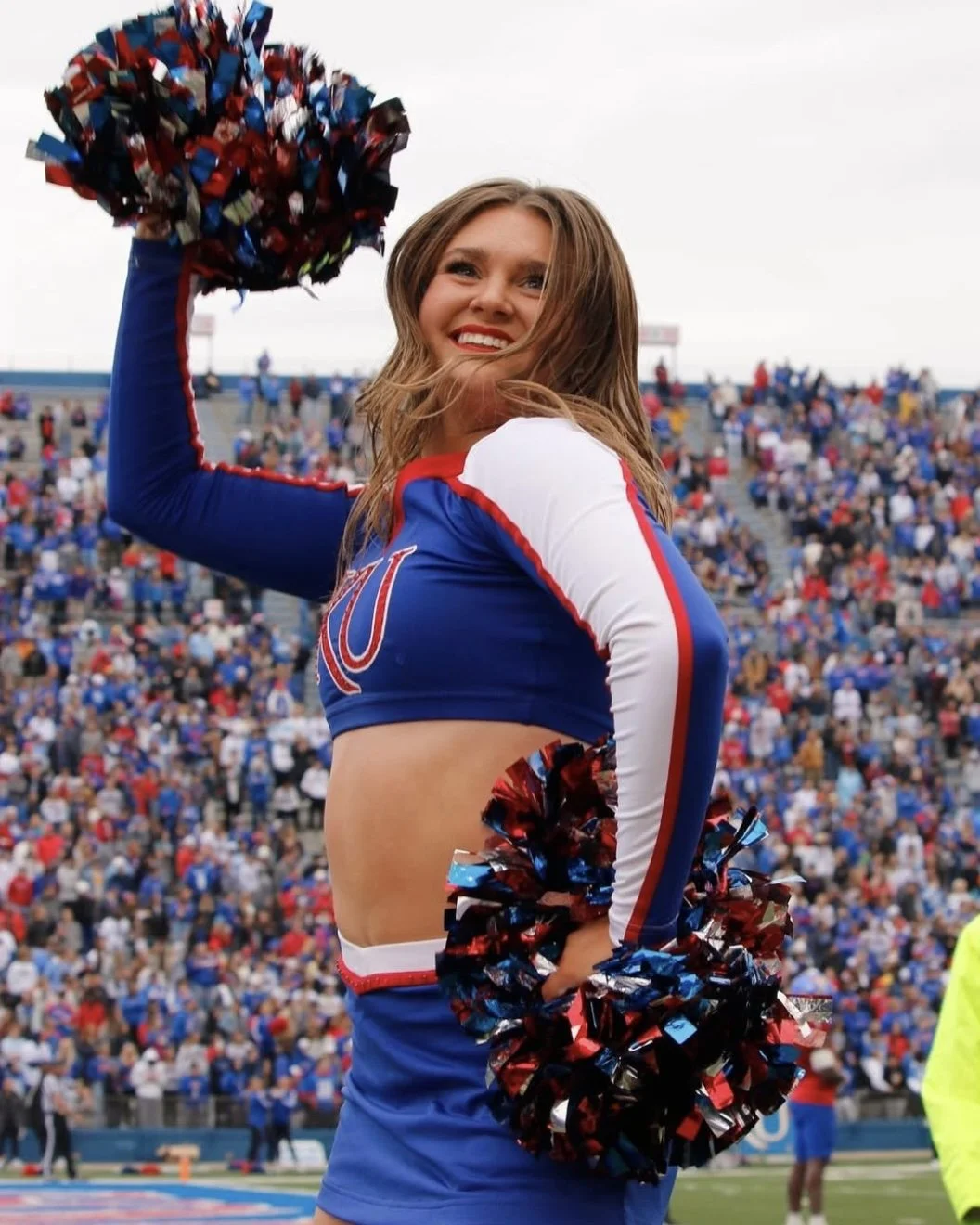 A young woman cheerleader in a blue and white uniform with red accents, holding red, blue, and black pom-poms at a sporting event with a crowd in the background.