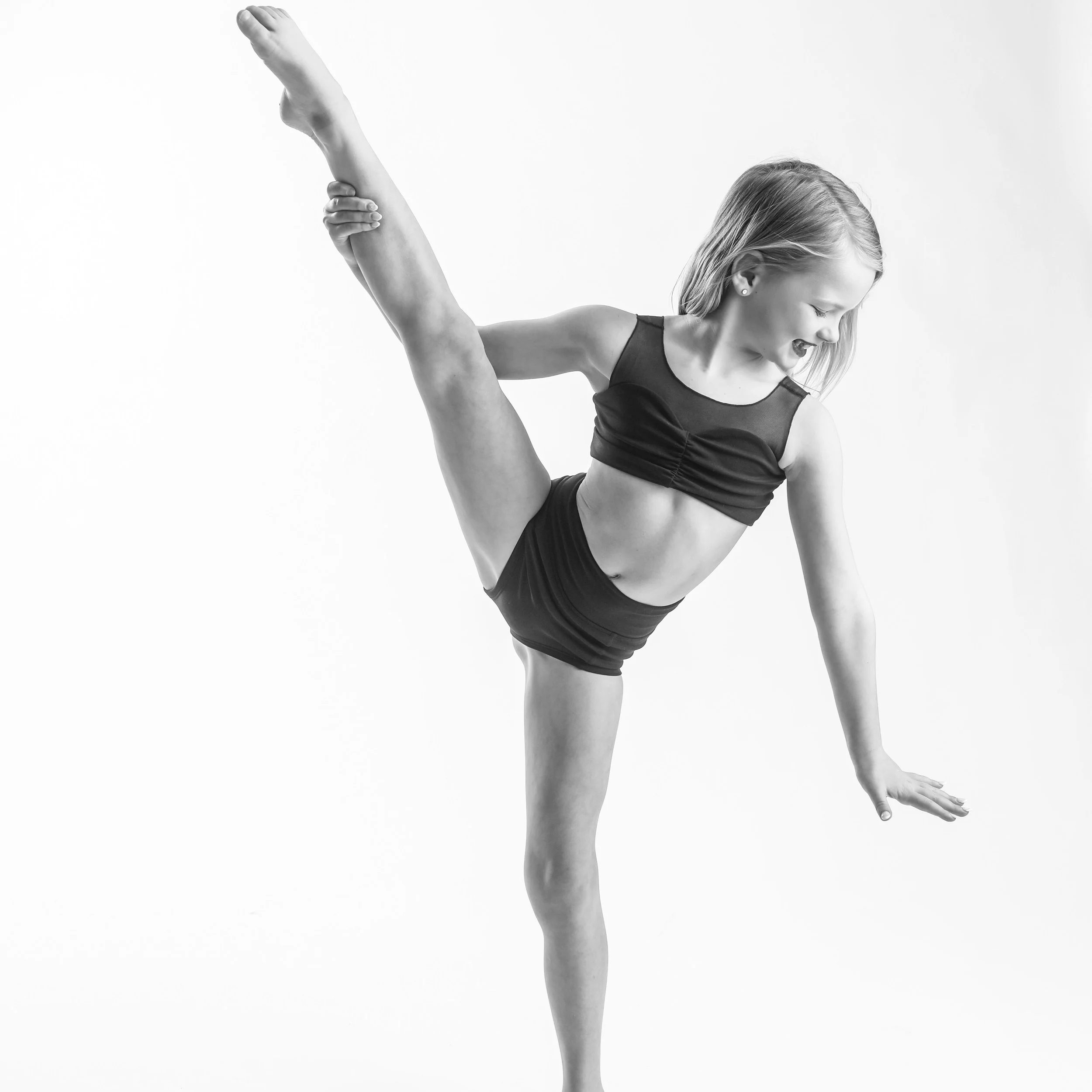 A young girl with blonde hair dressed in a black sports bra and shorts performing a yoga pose with one leg raised high and holding it with one hand, smiling and balancing on her other leg.