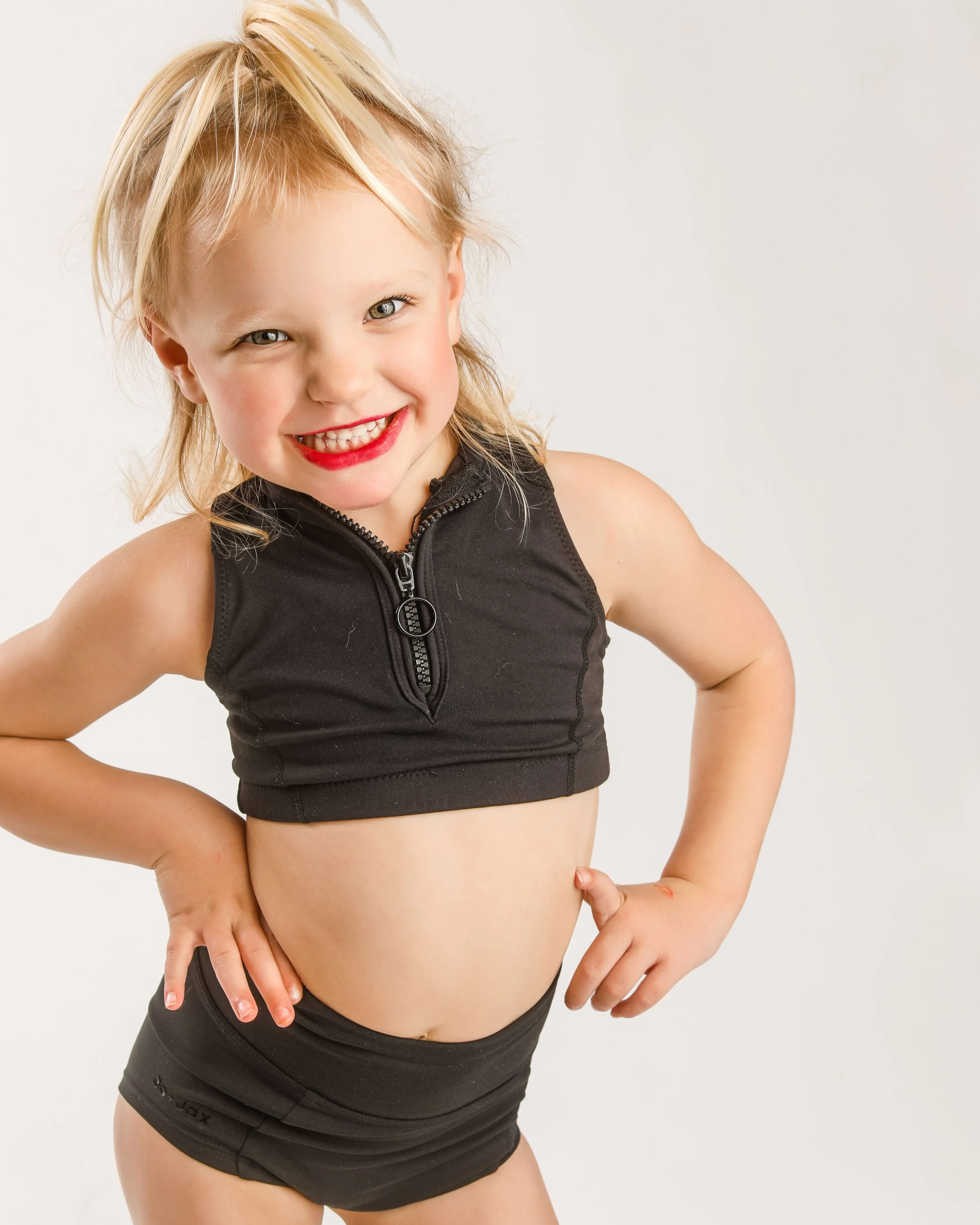 Smiling young girl with blonde hair, wearing a black sleeveless top and black shorts, posing with her hands on her hips against a white background.