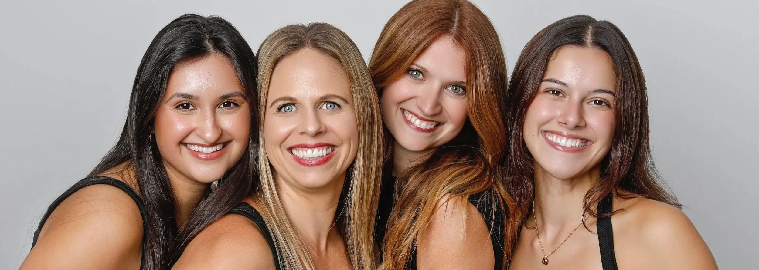 Four smiling women with different hair colors and styles, close together against a gray background.