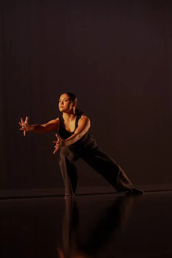 A woman in black athletic clothing performing a dance or movement pose on a dark stage with minimal lighting.