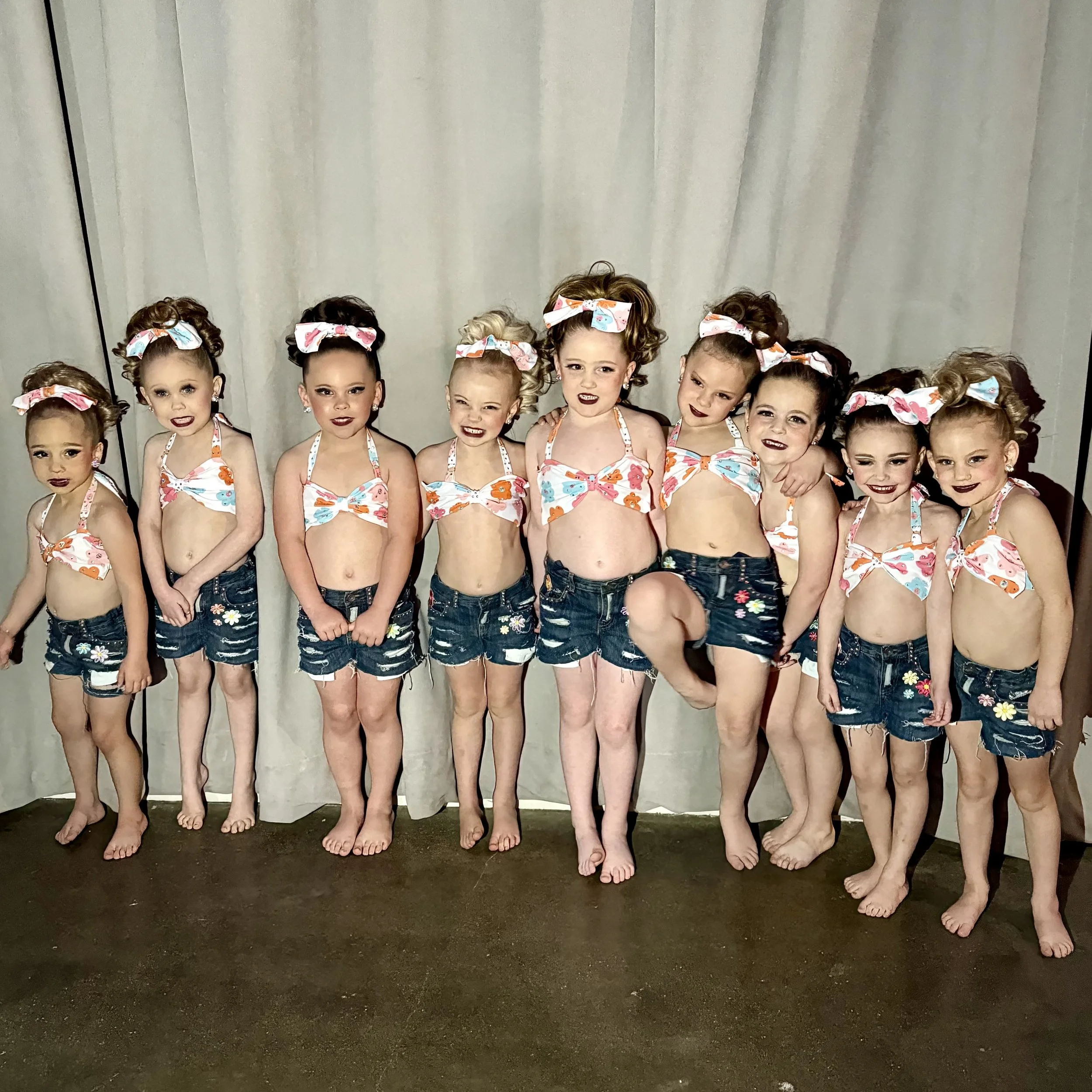 Group of ten young girls dressed in matching outfits with floral tops, denim shorts, and headbands standing in front of a curtain.
