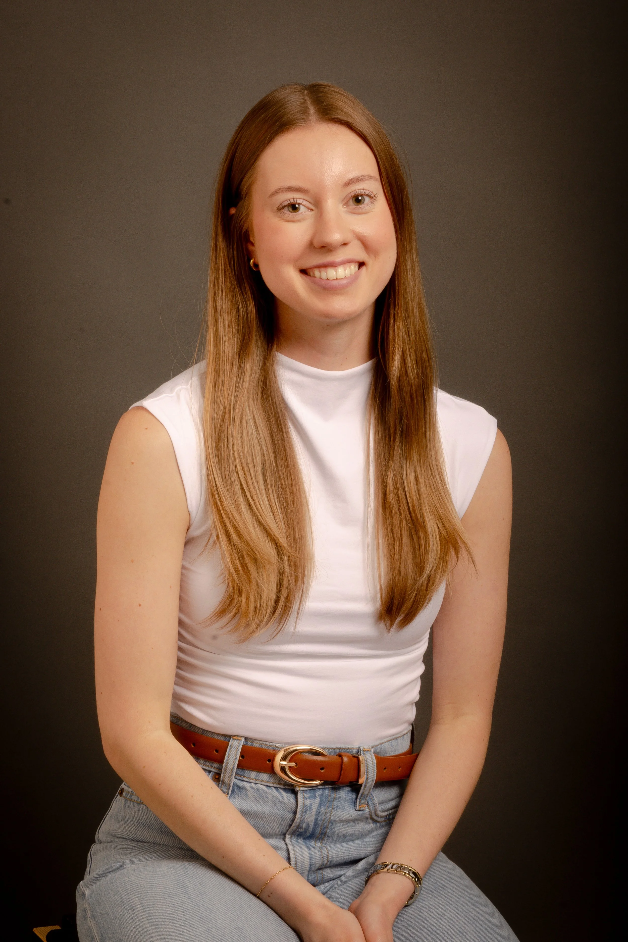 A young woman with long red hair, wearing a white sleeveless top, blue jeans, a brown belt, and gold jewelry, smiling and sitting against a dark background.