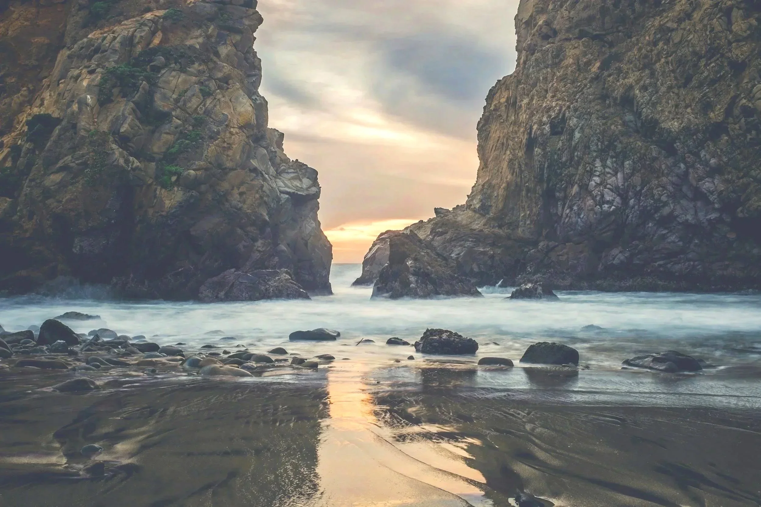 A rugged coastal scene at sunset with large rocky cliffs on both sides framing the water. The waves gently hit a pebbly shoreline in the foreground, and the sky features a mix of clouds with a hint of sunlight near the horizon.